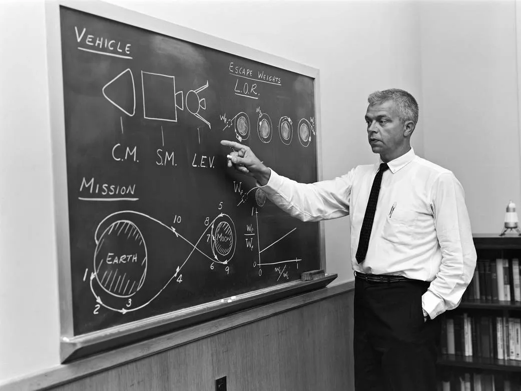 John Houbolt explains lunar-orbit rendezvous beside a chalkboard of spacecraft diagrams and calculations.