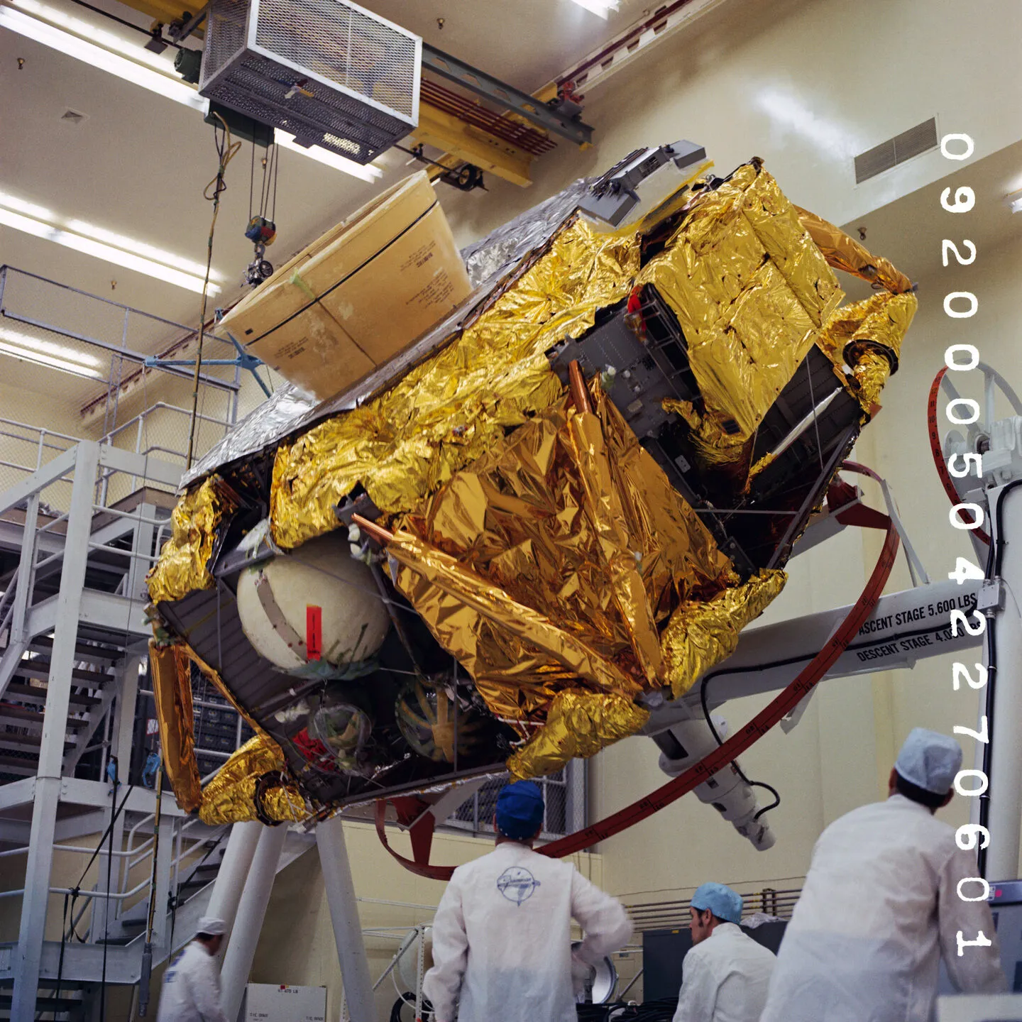 Technicians in white coats watching a lunar module descent stage, wrapped in gold thermal foil, being lowered onto a fixture on the factory floor at Grumman