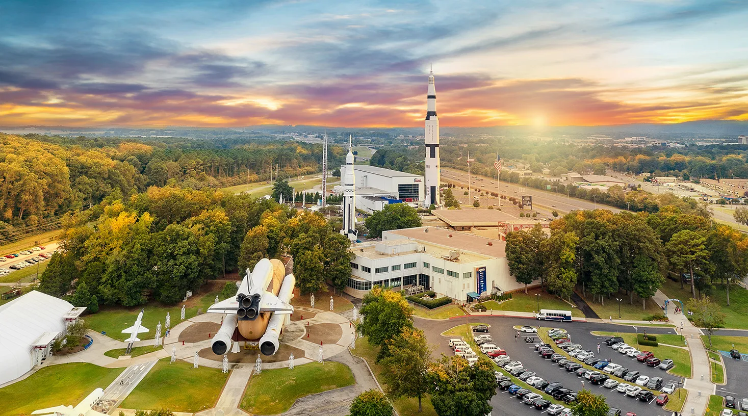 Aerial view of the U.S. Space and Rocket Center campus at sunset — the standing Saturn V, the Pathfinder shuttle stack, museum halls and Rocket Park among autumn trees