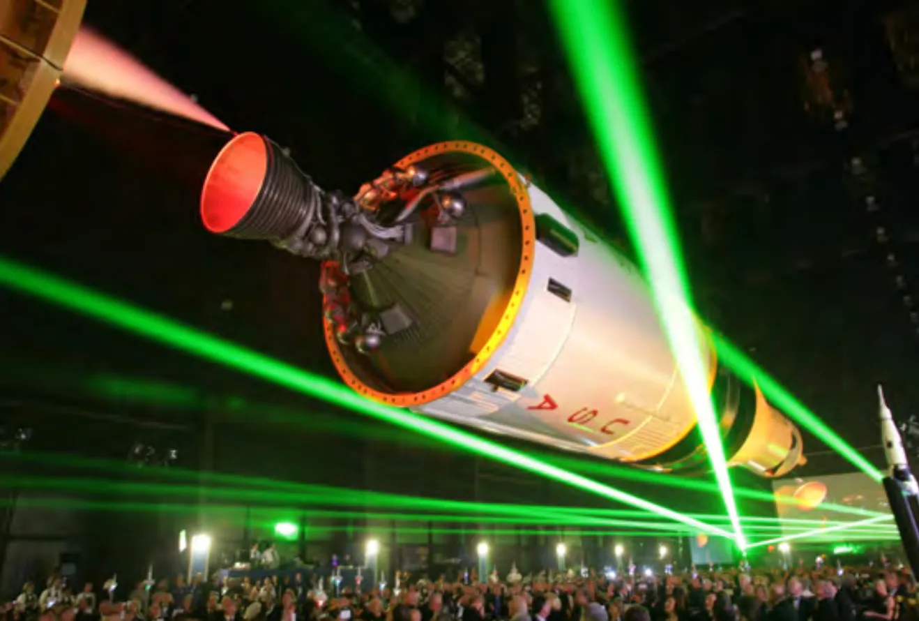 A crowd gathered beneath the Saturn V engine stage under a sweep of laser light